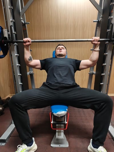 An adult male is observed performing a weightlifting exercise in a gym located in the city of Im. Gastello, Russia. The individual, dressed in a black short-sleeved athletic shirt, black athletic pants, and white and yellow athletic shoes, is lying supine on a blue-padded weight bench. The bench's blue upholstery is torn, revealing white material underneath. He is gripping an unweighted barbell, positioned at the top of a repetition within a grey metal Smith machine. His arms are extended upwards, and his legs are spread wide with feet on the floor. A dark tattoo, resembling a stylized cross or shield, is visible on his left bicep. A silver-colored beaded bracelet is worn on his right wrist. The background features light wooden paneling. The floor is covered with dark reddish-brown rubber gym matting, and a partial view of a dark kettlebell is visible on the floor in the lower-left background.