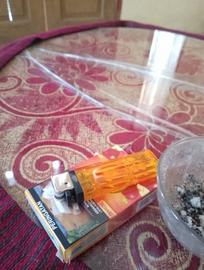 The image shows a close-up of a cigarette pack, a lighter, and an ashtray on a table with a patterned tablecloth and a clear plastic cover. The cigarette pack is red and yellow with text in Indonesian that says "PERINGATAN" (Warning) and "DIARUM" (a brand name). A silver and black lighter with an orange plastic casing is placed on top of the cigarette pack. The ashtray is a round, gray glass bowl filled with cigarette butts and ash. The background appears to be a wooden surface, possibly a door or furniture. The lighting suggests an indoor setting during the day. The location is specified as Sukamanah, Indonesia.
