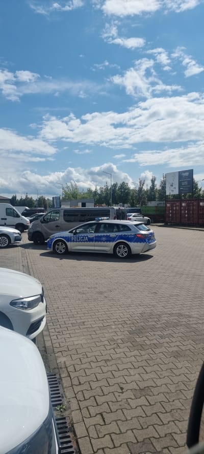 In Mroków, Poland, on a bright, cloudy day, a police car with the word "POLICJA" clearly visible on its side is parked on a paved lot. The police car is a silver station wagon with blue and white markings. Several other vehicles, including vans and cars, are also present in the lot. In the background, there are trees, buildings, and shipping containers. The sky is a vibrant blue with scattered white clouds. The ground is covered in interlocking pavers.