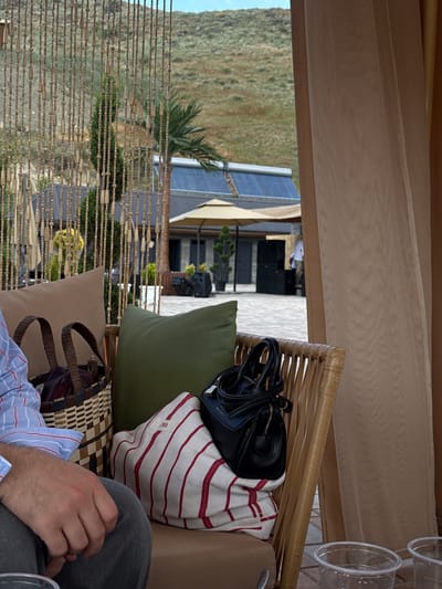 The image depicts an outdoor seating area at a venue in Ptghni, Armenia. In the foreground, a person wearing a blue and white striped shirt and grey pants is seated on a chair, with their right hand resting on their lap. Next to them are two pillows, one green and one tan, and a woven basket with a red interior. A white fabric bag with red stripes and a black handbag are also visible on the seating. In the background, a beaded curtain separates the seating area from a courtyard. Beyond the courtyard, there are buildings with dark roofs, awnings, and trees. A person in a white shirt and pants can be seen standing near the buildings. The environment appears to be a resort or event space, with a hilly, vegetated landscape visible in the distance under a bright sky.