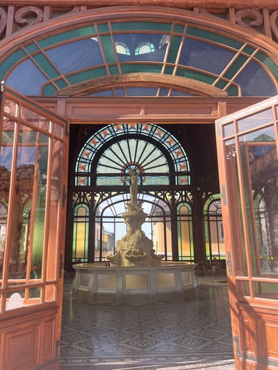 The image depicts the interior of the thermal baths in Évian-les-Bains, France, specifically the entrance hall leading to the central fountain area. The hall features ornate wooden architecture with large, open doorways and extensive use of stained glass windows. The focal point is a large, multi-tiered stone fountain with a statue at its apex, illuminated by natural light streaming through the colorful stained glass. The floor is covered with a decorative tiled pattern. In the background, beyond the fountain, two individuals are seated on chairs, seemingly resting. The overall impression is one of grandeur and historical significance, characteristic of Belle Époque architecture.