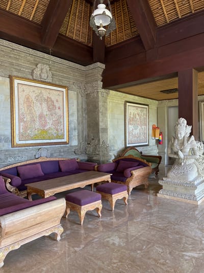 The image depicts an interior scene in Kedewatan, Indonesia, featuring ornately decorated furniture, artwork, and architectural details. The furniture set includes a long, upholstered sofa with purple cushions, accompanied by matching chairs and tables with ornate, gold-colored trim. Two framed paintings hang on the wall, depicting intricate, detailed scenes. A large white statue stands on a platform in the background. The room's architectural features include a thatched roof, dark wooden beams, and carved stone walls. A chandelier hangs from the ceiling. The floor is covered in marble tiles. There are no people visible in the scene.