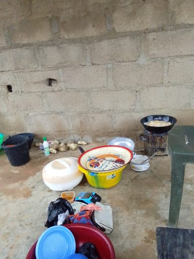 This image depicts an outdoor scene in Zanzi, Nigeria, likely a domestic setting or a makeshift kitchen area. The background is a rough-hewn brick wall, showing signs of weathering and age. In the foreground, various objects are scattered on a concrete floor.

To the left, there are two black buckets, one partially filled. Next to them, a green plastic bucket is visible. A white bottle with a green cap, possibly containing liquid, stands upright near a pile of what appear to be root vegetables, possibly yams.

Dominating the center of the image is a large, round, cream-colored insulated food container with a handle. Adjacent to this is a colorful, shallow bowl with a red rim, featuring a painted design of birds and flowers. Inside this bowl, cutlery is visible. A smaller white pot with two handles and a spoon is placed to the right of the bowl.

Further to the right, a small metal stove is in operation, with a black frying pan on top. The pan contains what appears to be food being fried in oil. Next to the stove, a dark green plastic stool stands.

In the lower part of the image, several items are grouped together. A black plastic bag lies on the ground, along with a blue electronic device that resembles a point-of-sale terminal. Beneath these, a red basin contains blue plastic plates and some black plastic bags. A folded piece of fabric with a red, blue, and white pattern is also visible.

The overall impression is one of everyday life, with cooking activities and domestic items set out in an open-air environment. The lighting suggests it is daytime.