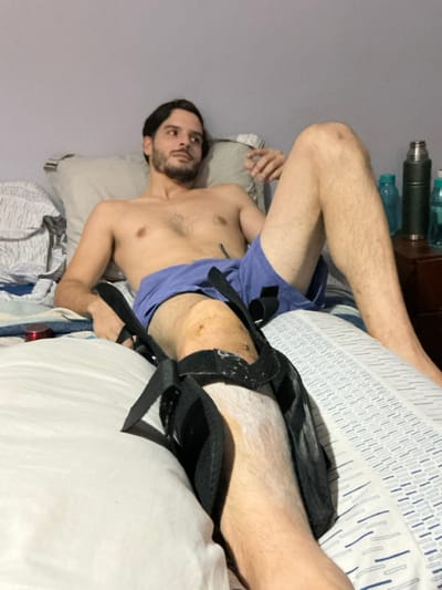 A man with a beard lies on a bed with his bare chest visible. He's wearing blue shorts. His right leg is in a black brace. The bed has a white and blue patterned pillow. There are water bottles on a table behind him. The wall is a light purple color.
