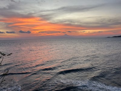 This image depicts a sunset over the sea in Desa Batu Layar Barat, Indonesia. The sky occupies the upper portion, displaying vibrant horizontal streaks of orange, red, and pink clouds near the horizon, fading into lighter grey and white towards the zenith. The lower two-thirds of the frame show a wide expanse of ocean, with the water surface reflecting the sky's colors in shades of dark blue, grey, and subtle pink. Gentle waves with visible white foam crests are prominent in the immediate foreground, indicating proximity to a shoreline or elevated vantage point. In the distant left, a distinct, symmetrical conical silhouette of a mountain or volcano rises above the horizon. A distant, low-lying landmass is visible on the far right horizon. Two small, dark, elongated objects, consistent with small boats, are present on the water in the mid-distance. A dark, leafy branch extends into the frame from the bottom-left corner. No human figures are visible.