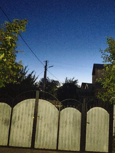 The image depicts a scene at dusk in the city of Ocnița, Moldova. In the foreground, a light-colored, arched wooden fence spans the width of the frame, with visible vertical planks and dark metal supports. A yellow padlock is attached to the central vertical support of the fence. Behind the fence, silhouetted trees and vegetation form a dark line across the midground. A wooden utility pole with electrical wires is visible on the right side of the frame, extending upwards into the sky. Further in the background, two dark-roofed structures are visible, one with a distinctive triangular window illuminated from within, forming the shape of an "A". The sky above is a gradient of deep blue at the top, transitioning to a lighter blue near the horizon, indicative of twilight. The overall atmosphere is calm and serene.
