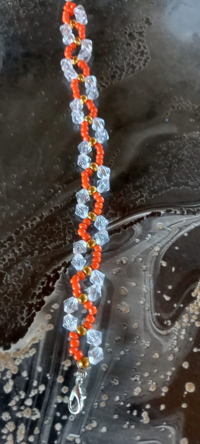 A beaded bracelet with clear faceted beads and orange round beads, interspersed with small gold beads, is laid out vertically on a dark, glossy surface with a swirling, marbled pattern. A silver lobster clasp is attached to the bottom end of the bracelet. The background surface appears to be an artificial material, possibly a countertop or table. No people are present in the image, and no context of a city or specific location is provided.
