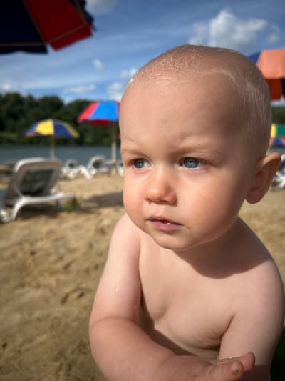 The media file shows a close-up, tilted shot of a baby's face and upper body. The baby has fair skin and light blonde hair. Their eyes are open and looking towards the viewer, and their lips are slightly parted. The baby's skin appears wet in places, and there is a small smudge of dirt on their finger. The background suggests an outdoor setting, with sand visible in the foreground and blurred elements of beach umbrellas and trees in the distance. The sky, also visible, appears blue with some clouds. The lighting indicates it is daytime, likely sunny. There is no visible text in the media file.