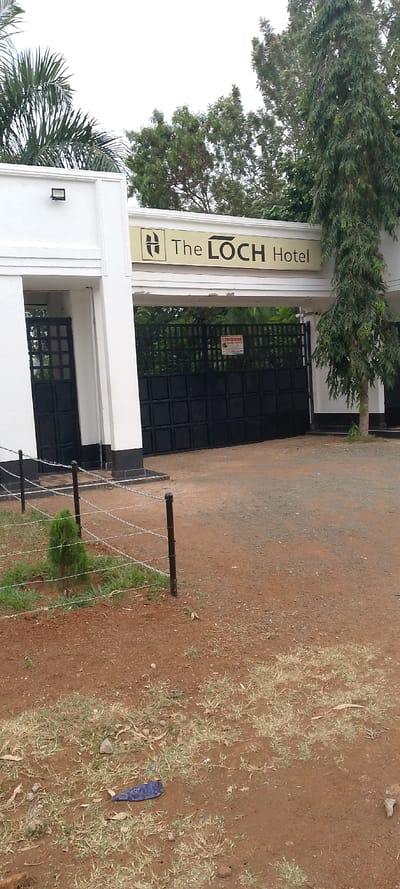 The image displays the outdoor entrance to "The LOCH Hotel" in South Sakwa ward, Kenya, under an overcast sky. The hotel entrance features a white architectural structure framing a closed, dark black metal vehicle gate with square panels. To the left of this main gate, a smaller, open dark black metal pedestrian gate is visible within the white framework. A light-colored sign with the dark text "The LOCH Hotel" and a stylized 'H' logo is mounted above the gates. A red and white warning sign, partially legible as "LINDUM", is affixed to the main vehicle gate.
On the left side of the entrance structure, a white wall section features a black rectangular outdoor light fixture, with large green palm fronds visible above. To the right of the entrance, a tall, slender tree with dark green, narrow leaves stands immediately adjacent to the white structure. The ground in the foreground is reddish-brown soil, mixed with sparse patches of green and dry grass and scattered small rocks. A crumpled blue plastic item lies on the soil in the lower-center-left. A short fence, composed of black metal posts and multiple strands of barbed wire, runs across the lower-left portion of the frame, enclosing a small, young green conifer-like bush. Various other green trees with dense foliage are visible behind and above the hotel entrance, suggesting a lush environment within. No people, specific actions, or interactions are depicted.