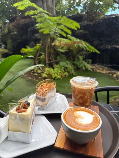 A close-up shot captures a tray holding two slices of cake and two beverages, set against a lush green outdoor environment with a rocky waterfall and lush foliage in the background. The tray, dark brown and round, is placed on a checkered surface. In the foreground, a white ceramic mug with latte art in the shape of a heart sits on a light brown wooden saucer. To its right, a clear plastic cup contains a cold coffee drink with swirling caramel-colored milk.
The cakes are presented on white plates. The slice on the left is a layered cake with a pale yellow frosting, adorned with nuts and a blueberry. The slice on the right is a taller, cylindrical cake, also layered and topped with what appears to be toasted almonds and herbs.
The background suggests a tranquil garden setting, possibly a cafe or restaurant with an outdoor seating area. The vibrant green of the artificial turf and the natural greenery, including ferns and trees, creates a visually appealing contrast with the food and drinks. The soft focus in the background draws attention to the foreground subjects. This scene likely depicts a relaxing break in Ban Na Chom Thian, Thailand, a location known for its coastal beauty and growing cafe culture.