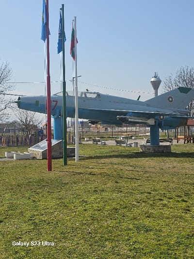 The image depicts an outdoor public park or memorial area in Dobroslavtsi, Bulgaria, under a clear blue sky. The central subject is a static, decommissioned MiG-21 fighter jet, light grey-blue in color, mounted on a blue cylindrical pedestal with a dark grey base. The aircraft features a prominent red number "7" on its nose, a Bulgarian Air Force roundel on its tail, and two dummy missiles attached under its wings.
In the foreground, a grassy expanse with varying shades of green and some drier patches extends towards the viewer. Directly in front of the aircraft, three flagpoles are erected: a red pole flying a blue flag, a green pole flying another blue flag, and a white pole displaying the Bulgarian national flag (white, green, and red horizontal stripes). To the left of the aircraft's pedestal, a small memorial or monument with a dark base and lighter top is visible.
Beyond the aircraft, the environment includes multiple light-colored concrete park benches, concrete planters, and a children's play structure with colorful vertical bars. Bare tree branches are visible on the left and right edges of the frame. In the distant background, a tall, slender water or communication tower with an antenna array stands prominently on the right side of the horizon. No people are present in the scene. The lighting indicates a sunny day.
