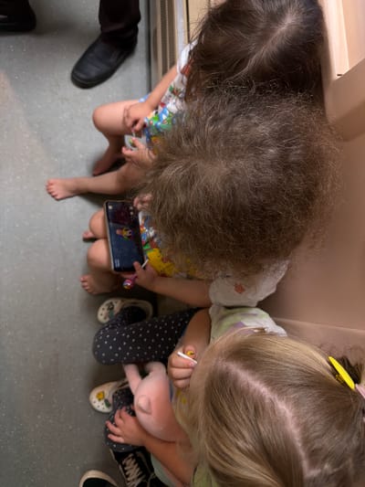The image shows a top-down view of four children sitting next to each other, possibly on a bench or floor, indoors. The environment appears to be a public space, suggested by the gray speckled floor and a vertical metal grate visible in the background. An adult's dark-colored shoes are partially visible in the upper left corner.
The children are positioned from top to bottom in the frame. The topmost child is largely obscured by the child below. The second child from the top has dark, curly hair and is holding a smartphone, which displays a colorful image of a person. This child is also holding a lollipop with a pink and blue swirl. Their legs are bent at the knees, and their feet are bare.
Below this child is another child wearing a dark, polka-dotted outfit and white clogs. This child is holding a pink plush toy. Their legs are also bent.
The child at the bottom of the frame has blonde hair with a yellow hair clip and is wearing light-colored clothing. This child is also holding a small white object, possibly a piece of candy or a toy.
The overall scene depicts children engaged in activities while waiting or resting in a communal area. The location is identified as Zykovo, Russia.