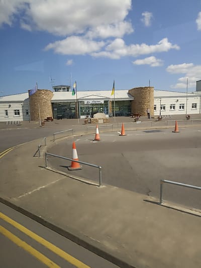 This is a daytime, outdoor, medium-distance, eye-level shot of the Donegal Airport terminal building, located in Carrickfinn, Ireland. The building is modern with a white, sloped roof and a facade featuring large glass windows and two circular stone towers. An EU flag and a green and yellow flag are flying from poles in front of the building. A man is standing near the entrance. In the foreground, there is an asphalt road with yellow double lines, and a sidewalk with metal railings. Several orange traffic cones are placed on the road and sidewalk, suggesting ongoing work or traffic management. The sky is blue with scattered white clouds.