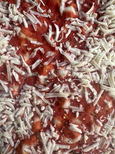 An overhead close-up shot shows a raw, uncooked pizza covered with shredded white cheese and a vibrant red tomato sauce. The cheese is layered unevenly over the sauce, with some areas showing more sauce and others more cheese. Small flecks of herbs, likely oregano or basil, are visible mixed with the cheese and sauce. The pizza appears to be on a flat surface, possibly a baking sheet or pizza stone, as suggested by subtle textures in the background beneath the pizza. There are no people or specific environmental features of Golyam Chardak, Bulgaria visible in this image.
