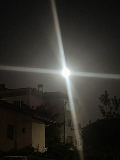The image captures a nighttime scene in the city of Peshtera, Bulgaria. A bright, ethereal light source, possibly the moon or a strong artificial light, dominates the sky with a lens flare effect, creating a prominent cross-shaped glare across the frame. Below this celestial display, the silhouettes of buildings are visible. A multi-story building with visible windows and balconies is in the mid-ground, partially obscured by trees. In the foreground, another building with a dark roof and windows is also present. The overall atmosphere is dark and moody, characteristic of a nocturnal urban environment.