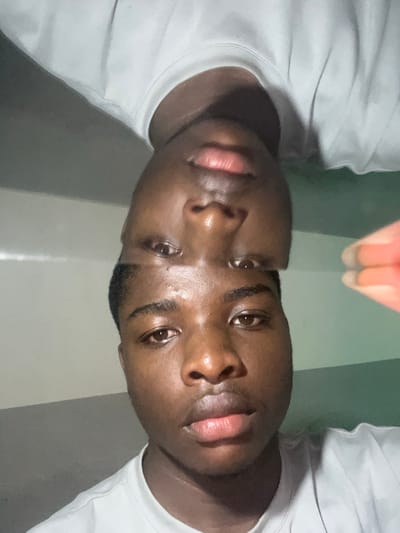 The scene depicts a young adult male located in the city of Shen, Nigeria. The individual has dark skin, short dark hair, and brown eyes, and is wearing a white crew-neck t-shirt. The image features a prominent split composition: the lower section displays his face in an upright orientation, while the upper section shows an inverted, partial reflection of his face. Both facial representations are directed forward, engaging the viewer. A reddish-orange object, consistent with a finger or part of a device, is partially visible in the upper right quadrant. The background consists of a plain wall, light grey on the left and transitioning to a greenish-grey hue on the right. No other individuals or actions are discernible within the frame.