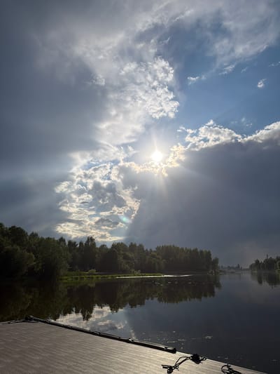 The image shows a body of water, likely a river or lake, with reflections of the sky and trees. On the left, a wooden platform is visible in the foreground. The sky is dramatic, with a mix of dark clouds and patches of bright sunlight breaking through, creating visible rays. Trees line the far bank of the water, creating a natural landscape. In the distance, along the shoreline, there are some structures, possibly houses or other buildings, and more trees. The overall scene depicts a natural environment with a hint of civilization, possibly located in Shchelkovo, Russia, given the context. No people or specific actions are depicted.