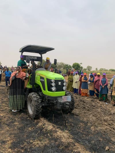 The image shows a bright green tractor with a black canopy parked in a field of burnt vegetation in Ringimawa, Nigeria. There are approximately 20 people in the scene. A woman in a green and black dress and a pink headscarf is speaking into a microphone in front of the tractor. Another woman in a blue shirt and a purple hat is standing next to her. A man in a yellow hard hat is sitting in the tractor’s cab. There are men and women, some wearing colorful hats, gathered behind the tractor. They appear to be listening to the woman who is speaking. The tractor is labeled "WOFAN" on the sides and the cab has the same label on the back. The ground is mostly bare, with burnt vegetation, and mud. The sky is overcast with clouds. The scene was captured at the coordinates 11.666 latitude and 8.415 longitude, a location within the city of Ringimawa in Nigeria.