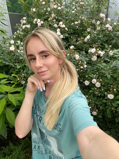 A young woman with blonde hair and light skin is taking a selfie outdoors. She is wearing a teal t-shirt. Her left arm is bent and her hand is resting on her chin, with light purple nail polish visible. She has light brown eyes and is wearing subtle makeup. Behind her is a large bush with numerous small, pale pink and white roses in bloom. Green foliage surrounds the roses and her. To the left of the frame, there are also some larger green leaves from another plant. In the background, a green chain-link fence and a grey metal structure are partially visible. The overall impression is that of a warm, outdoor setting, possibly a garden. This scene is located in the city of Yasnoe, Russia.
