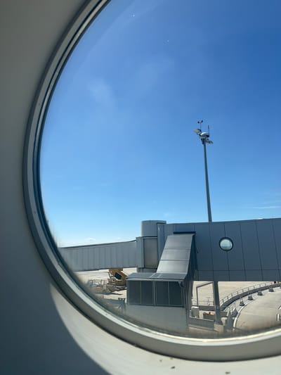 The image shows a view from an airplane window at Schwechat, Austria. The sky is clear and blue, suggesting good weather. Outside the window, we can see parts of an airport terminal building and a jet bridge. A weather station, possibly an anemometer, is visible on a tall pole extending into the sky. The sun is shining, casting shadows on the ground, indicating it is daytime. The perspective suggests the plane is parked at a gate. There are no people visible in the image. The scene is an active airport environment.
