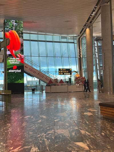 The image depicts the interior of a modern airport terminal, likely at Gardermoen, Norway, given the signage. The space features a high ceiling with a grid of ceiling tiles and large, concrete support columns. A massive floor-to-ceiling window dominates the background, offering a view of the sky and possibly the exterior of the airport.

On the left side of the image, a large LED screen displays an advertisement. The advertisement features a close-up of bright orange headphones against a backdrop of greenery, with text that reads "Livet blir bedre med lydbøker" and the Storytel logo.

A prominent escalator is visible, ascending from the lower level. A red directional sign with white text on the escalator reads "← Italian - PIZZA - Pasta".

Further into the terminal, a series of yellow directional signs indicate gates and toilets. One sign reads "Gates E,F,G" with an arrow pointing to the right, and below it, "Utgang Gates A,B,C,D" with a right arrow. Another sign indicates "Toalett Toilets" with circular icons.

In the mid-ground, a retail kiosk is set up, featuring a display of chocolates. The name "MENDEL'S" is visible on the kiosk.

Several people are present in the scene. Two individuals are walking together in the mid-ground, one in a dark t-shirt and the other in a black outfit. To the right, a person in a beige outfit is walking with a suitcase. Another person is seen standing near the base of the escalator.

The floor of the terminal is made of large, polished marble tiles with a grey and white veined pattern, reflecting the ambient light. In the foreground, a dark-colored bench with a wooden base is visible. The overall atmosphere appears to be bright and spacious, characteristic of a contemporary airport.