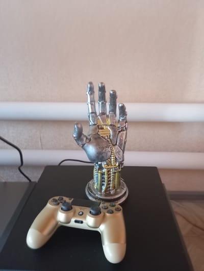 A metallic, prosthetic hand figurine stands on a black surface next to a gold video game controller. The hand is silver with gold mechanical detailing and is positioned upright. The video game controller is positioned horizontally in front of the figurine. The background consists of a textured, beige wall with two white horizontal pipes running across it. The scene appears to be indoors, possibly in a home entertainment area. There is no indication of the location being in the city of Высокая Гора, Russia.