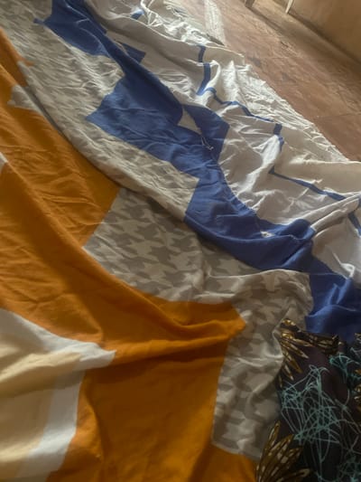 The image displays a close-up, angled view of a rumpled bed covered with a patterned duvet. The duvet features large blocks of solid colors: bright orange and royal blue, interspersed with sections of a grey houndstooth pattern and white fabric. Towards the lower right corner, a smaller portion of fabric with a dark blue base and a complex pattern of teal lines and brown floral-like shapes is visible, possibly a pillowcase or an additional blanket. The bedding is creased and folded, indicating it is not neatly arranged. The background, visible in the upper right quadrant, appears to be a wooden floor with distinct planks and possibly a wall or furniture made of wood. The lighting suggests an indoor environment, possibly a bedroom. The overall scene is domestic and appears to be a snapshot of a casually made bed. There are no people or animals visible in the image. The location is identified as the city of Kirsinga, Nigeria.