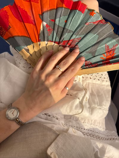 This is a close-up, over-the-shoulder shot of a person's arm and hand holding a partially opened fan with an artistic, floral and bird-like pattern in shades of orange, red, grey, and teal. The fan has a bamboo frame. The person is wearing a gold ring with a small diamond and a silver and gold-toned wristwatch. They are dressed in white clothing with intricate lace detailing and a light beige blazer or jacket. The setting appears to be indoors, possibly on a mode of transportation like a train or plane, given the dark background and the suggestion of a seat in the upper left corner. The lighting suggests it is daytime, with the sun casting a shadow on the person's hand and arm. The overall mood is calm and leisurely. There is no visible text in the image.