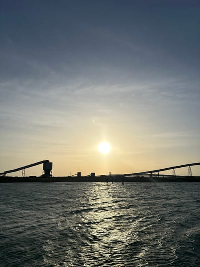 The image depicts a sunset over a body of water, likely a bay or ocean, with industrial structures silhouetted against the sky. The sun is low on the horizon, casting a warm, golden glow that reflects off the rippling water. The water appears choppy, with small waves creating a textured surface. In the background, a series of conveyor belts and silos are visible, suggesting an industrial port or mining facility. The sky is a gradient of blues and yellows, with thin clouds streaking across the upper half. The overall mood is serene and slightly industrial. The location is identified as Uribia, Colombia, a city known for its proximity to coal mining operations and the Caribbean coast, which aligns with the visual elements of the image.