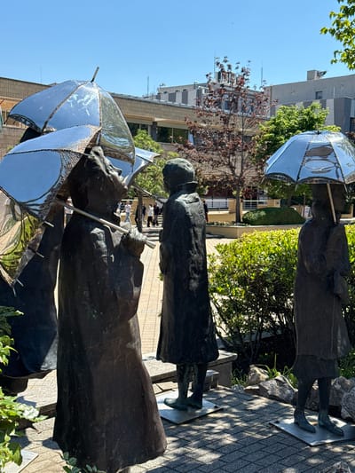 A bronze statue of three children under umbrellas stands on a tiled surface in an outdoor urban environment. The umbrellas have a reflective, possibly metallic, silver finish. The children are depicted in simple, draped clothing. The scene is set against a backdrop of greenery, trees, and urban buildings. The location is likely Siófok, Hungary.
