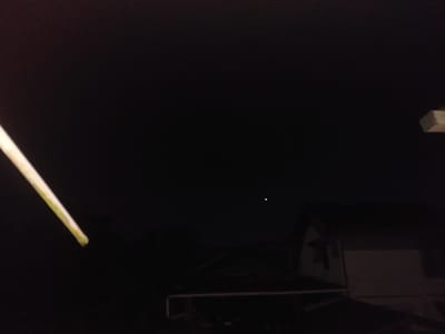 The image shows a dark, nocturnal scene. In the foreground on the left, a light-colored object, possibly a pole or a thick stick, is angled into the frame from the bottom left, its tip partially visible. The background is dominated by a black sky, punctuated by two small, bright dots of light, suggesting stars or celestial bodies. Below the sky, a silhouette of a building is visible, appearing to be a house with a sloped roof. A horizontal structure, possibly a fence or railing, is in front of the house. The overall setting appears to be outdoors, likely in a residential area, given the presence of the house. The time of day is clearly night. There is no visible text in the image. The mood is dark and quiet, with a focus on the vastness of the night sky and the subtle lights within it, contrasting with the grounded presence of the house. The location context of Jelutung, Indonesia, suggests a tropical or subtropical environment, though this is not directly discernible from the image itself.