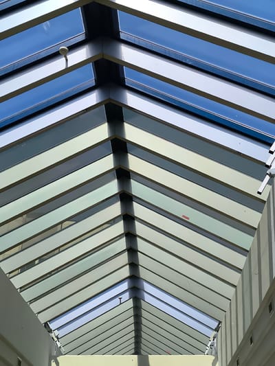 The image depicts an interior view looking up at a skylight structure, possibly in Schwechat, Austria. The skylight is composed of alternating panes of glass and solid, possibly metal, beams, creating a striped pattern. The panes alternate in color, suggesting different types of glass or light transmission properties. At the top of the structure, the sky is visible through the glass. A security camera is mounted on one of the beams. The architecture is modern, with clean lines and a symmetrical design. The walls are light-colored and appear to be part of an indoor space.