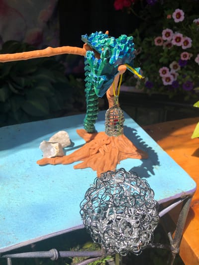 A 3D printed dragon sculpture, colored in shades of blue and green, hangs from an orange branch-like structure. Suspended below the dragon by a yellow string is a miniature silver birdcage containing a red object. The dragon and cage are perched on an orange, tree-stump-like base. Two small rocks are placed near the base. In the foreground, a spherical object made of intertwined silver wire rests on a light blue table. In the background, a dark green plant with lush leaves is visible, along with a bush adorned with pink and purple flowers. The scene is set outdoors under natural sunlight, casting shadows on the table. There are no people visible in the image. The location is an outdoor setting, possibly a garden or patio, in the city of Severn, Canada.
