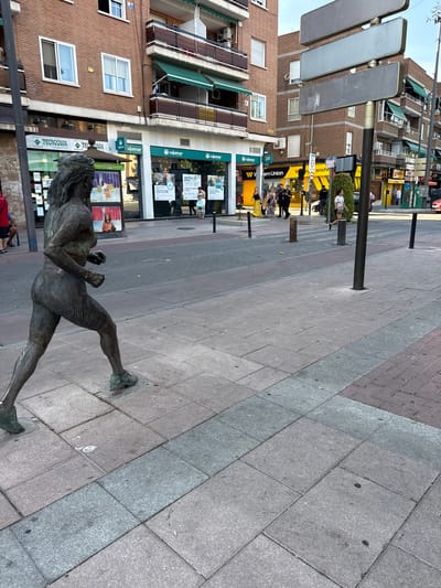 The image shows a bronze statue of a woman running, placed on a pedestrian walkway in the city of Getafe, Spain. The statue is positioned in the foreground on the left side of the frame, facing right. The background features buildings with red brick facades, shops including "Tecnocasa" and "Cajamar," and street signs. Several people are visible in the background, walking along the street and sidewalk. The environment appears to be an urban street scene with a clear sky. The lighting suggests it is daytime.