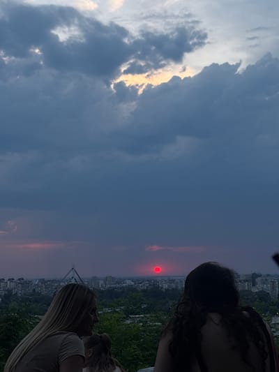 The image depicts two individuals from behind, facing a cityscape under a dramatic sky. The person on the left has long blonde hair and is wearing a light-colored shirt. The person on the right has long, dark, curly hair. They are seated on what appears to be an elevated viewpoint, with lush greenery and a city skyline below. The cityscape includes buildings and a distinctive structure, possibly a bridge or tower, in the distance.
The sky is filled with dark, ominous clouds, with a sliver of bright light peeking through near the top. Dominating the middle distance is a striking red sun, appearing as a distinct, spherical orb low on the horizon. The lower portion of the sky surrounding the sun is tinged with pink and purple hues, indicative of sunset or twilight.
The scene suggests a moment of observation, possibly contemplation, as the individuals take in the view of the city and the striking sunset. The presence of the cityscape and the distinct red sun points to Petrovaradin, Serbia, as the likely location. The overall atmosphere is moody and serene, with the vibrant red sun providing a focal point against the brooding sky.