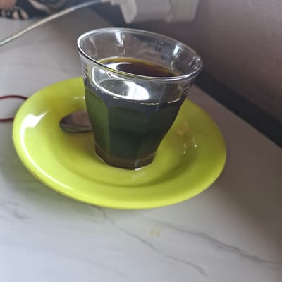 A close-up, high-angle shot shows a clear glass of dark liquid, likely coffee, sitting on a bright yellow saucer. A silver spoon is partially visible on the saucer, to the left of the glass. The background is blurred, showing a grey and white marbled surface and a dark, out-of-focus object above. The scene appears to be indoors, possibly in a cafe or home setting, in the city of Mesjid Tuha, Indonesia.