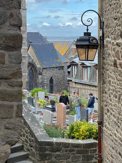 The image captures a view looking down into a cemetery in Le Mont-Saint-Michel, France. The cemetery is situated amongst ancient stone buildings with grey slate roofs, some showing patches of yellow lichen. A vintage black street lamp with a scrolled arm is affixed to a stone wall on the right side of the frame, its amber glass panels dimly lit. In the cemetery, gravestones of various sizes and materials, including granite and marble, are arranged amongst green foliage and red flowers. Three men are standing in the cemetery, appearing to be looking at gravestones or conversing. One man in a black sweatshirt stands with his back to the camera, holding a dark object. Another man in a green jacket and jeans stands to his left, while a third man in a blue jacket and denim shorts stands to his right. The background shows a blurred expanse of the town and distant landscape under a cloudy sky. The foreground features a textured stone wall and a set of stone steps leading down into the scene.