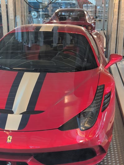 The image shows the front of a red Ferrari with racing stripes, likely at the Mercedes-Benz Museum in Stuttgart, Germany. A second, darker red car is visible in the background, also appearing to be a sports car, possibly a Ferrari as well, and is parked behind a black barrier. The cars are indoors, in a brightly lit environment that resembles a showroom or exhibition space, with reflective flooring and industrial-looking metal structures and signage in the background. There are no people visible in the image.