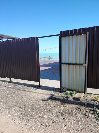 The image depicts an open corrugated metal gate in Toranghylyk, Kazakhstan, revealing a paved area with benches and a view of the sea under a clear blue sky. The gate itself is weathered, with rust stains streaking down its vertical panels. To the left, a solid dark brown corrugated metal fence extends out of frame, with a partial view of a covered structure at its far end. The ground in the foreground is covered with gravel and dirt. In the background, string lights are visible across the paved area, and the calm sea stretches to the horizon.