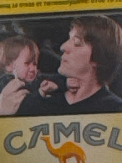 The image, rotated 90 degrees clockwise, depicts a close and dramatic interaction between two individuals, likely men or young men, in what appears to be an indoor or dimly lit setting.
The person positioned above has dark brown hair and is leaning downwards, looking intently at the other individual. Their right hand is shown in a clenched position or a fist, near the head of the person below. They are wearing dark clothing, and their facial expression appears focused or intense.
The second person, positioned below, has lighter, possibly reddish-brown hair and is looking upwards towards the first individual. Their face is visible, and a hand (belonging to either person) is near their chin or neck area. Their expression is engaged, though details are blurred.
The activity suggests a moment of physical tension, possibly a struggle or a staged performance, given the proximity and expressions.
To the right of the individuals, a distinct yellow-beige vertical band is visible, featuring the word "CAMEL" in large, stylized dark grey letters. Below this text, an orange graphic depicting a camel's head and hump is present. This indicates the image is likely printed on packaging, such as for Camel brand cigarettes. The background behind the figures is dark and lacks specific details. The overall image quality is somewhat blurry.