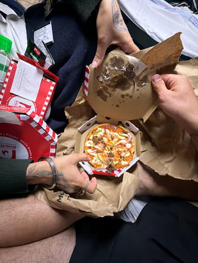 The image depicts a close-up, overhead view of a person's hands interacting with pizza boxes and food packaging. The scene appears to be indoors, with a person's legs, covered in hair, visible in the lower portion of the frame. The environment includes various pizza boxes, one of which is open revealing a small pizza topped with meat, vegetables, and cheese. There are also packets of spices and a plastic bottle with a green cap. The overall impression is one of a casual meal, likely in a domestic setting, possibly a bedroom given the presence of bedding-like material in the background. No specific landmarks or indicators of location within Surat, India, are present in the image.