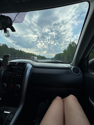 The view from inside a car shows a cloudy sky with trees lining the horizon as seen through the driver's side window. A semi-truck is visible on the road ahead. The dashboard includes climate controls with a digital display showing "21.0" with a "TEMP" label. Knees are visible on the passenger side. The odometer reads "53 000". The location is Beloborodovo, Russia.