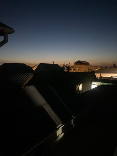 The image depicts a twilight scene over a residential area in Alamüdün, Kyrgyzstan. The sky is a deep gradient of dark blue to a faint orange near the horizon. Several buildings with dark, sloped roofs are visible in the foreground and mid-ground, some illuminated by interior lights. A downspout is visible on the left side of the frame, attached to a building. The overall impression is one of dusk settling over a quiet neighborhood.