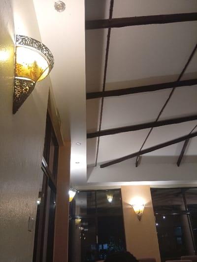 An interior view shows a building with a unique ceiling design featuring dark brown beams across a white surface. Ornate wall lamps provide a warm glow, enhancing the ambiance. The space is illuminated with natural light coming through expansive windows that overlook an outdoor setting. The building exhibits a blend of architectural elements, creating a cozy atmosphere.