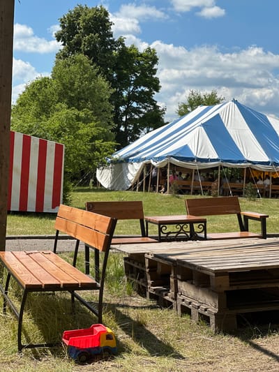The image depicts an outdoor scene with a large blue and white striped tent in the background, under which people are seated on benches. In the foreground, there are several wooden benches and tables, some constructed from repurposed wooden pallets. To the left of the scene, a red and white striped partition stands vertically. A small red, yellow, and blue toy truck rests on the grass near one of the benches. The environment appears to be a grassy area with trees in the background, under a partly cloudy sky. The location is identified as Завалочыцкі сельскі Савет, Belarus.