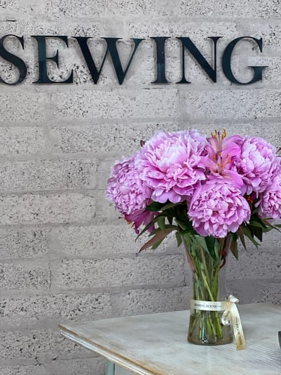 The image shows a bouquet of pink peonies in a glass vase, placed on a light-colored wooden surface. A white ribbon with "SEWING ROOM" printed in black text is tied around the vase. To the left of the vase, large, dark, three-dimensional letters spell out "SEWING" vertically on a textured, grey brick-like wall. The wall appears to be an interior wall, possibly in a home or commercial space. The lighting suggests an indoor setting, possibly during daylight hours. The overall impression is one of a decorative arrangement, likely in a space related to sewing or crafting, given the text on the ribbon and the wall letters. There are no people visible in the image. The location is specified as Donegal, Ireland, but there are no specific visual cues in the image to confirm this without external information.
