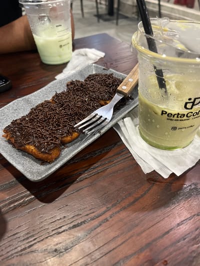 The image shows a close-up of a wooden table with food and drinks. In the foreground, a grey rectangular plate holds three pieces of fried banana topped with chocolate sprinkles. A silver fork with a wooden handle is placed on the plate. To the right of the plate is a clear plastic cup with a green beverage, likely matcha, and a black straw. The cup has condensation on the outside and a logo that reads "Perta Coffee". A white paper napkin is tucked under the cup. In the background, partially visible on the left, is another clear plastic cup containing a pale green beverage. A person's arm and shoulder are visible behind this cup. The table surface appears to be made of dark, polished wood with prominent grain. The setting is indoors, with the background showing out-of-focus shapes that suggest a cafe or restaurant environment. The presence of "Perta Coffee" on the cup indicates the location is likely a Perta Coffee outlet. While the prompt specifies the city of Meunasahceubrek, Indonesia, this detail cannot be independently verified from the visual information alone.
