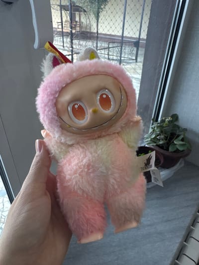 A human hand, displaying light-colored nail polish, holds a "The Monsters" Labubu plush toy indoors in Apshak, Uzbekistan. The toy features a fuzzy, ombre body transitioning from pink to yellow, a light-toned plastic face with large orange eyes, a small yellow nose, and a wide black smile revealing multiple white teeth. A yellow fabric tag with "THE MONSTERS" printed in red is visible on the toy's head. The background consists of a window offering a view to an outdoor area with paved ground, a dark metal grid fence, green foliage, and a distant wooden arched structure. To the right of the toy, a potted plant with green leaves rests on a surface, with a white grate or vent partially visible at the bottom right.