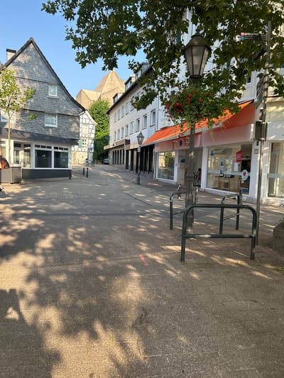 The image depicts a street scene in Velbert, Germany, likely a commercial area with shops lining both sides. The sun casts dappled shadows from trees onto the paved pedestrian walkway, creating a dappled light effect.

On the left, a two-story building with a dark slate-tiled roof and white-framed windows is visible. Adjacent to it is a building with half-timbered architectural elements. Further down the street, a series of modern buildings with large glass storefronts and orange awnings are present. One storefront displays the text "WBS TRAINING." Inside, a person is seated at a counter with stools.

On the right side of the street, more modern buildings with windows and signage are visible. A black lamppost with a hanging planter filled with red flowers stands near the foreground. A metal bike rack is positioned on the sidewalk. A street signpost with directional information, including distances to "Velbert" and other locations, is also present. In the background, a large, distinctive pyramidal concrete structure is visible. The sky is clear and blue.