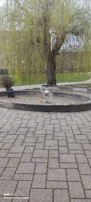 The scene is located in Constance, Germany, depicting an outdoor area under an overcast sky. The foreground is composed of a grey paver pathway laid in an interlocking basket-weave pattern. In the mid-ground, a circular planter constructed from dark, weathered timber or concrete contains light-colored sand. Centrally positioned within this planter is a silver-toned metal sculpture depicting a sphere encased by two intersecting elliptical rings. Directly behind the planter, a large weeping willow tree (Salix babylonica) with a thick, dark trunk exhibits abundant light green, newly unfurled foliage, characteristic of early spring. To the left of the planter, a smaller, dense bush with dark leaves is visible. The background consists of a grassy embankment and indistinct architectural structures partially visible through the tree's canopy. No individuals, animals, or observable actions or interactions are present in the image.